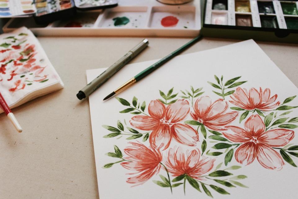 ภาพวาดดอกไม้สีน้ำโทนส้มชมพูบนกระดาษ พร้อมพู่กันและจานสี